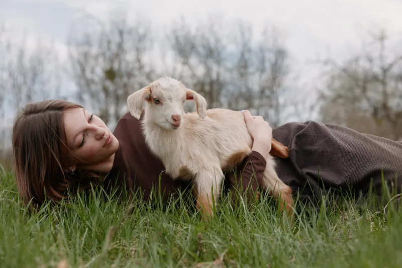 woman outdoors goat kid lifestyle nature grass smile, cheerful candid moment with a relaxed pose, sunlit scene and natural colors capturing a playful bond in nature