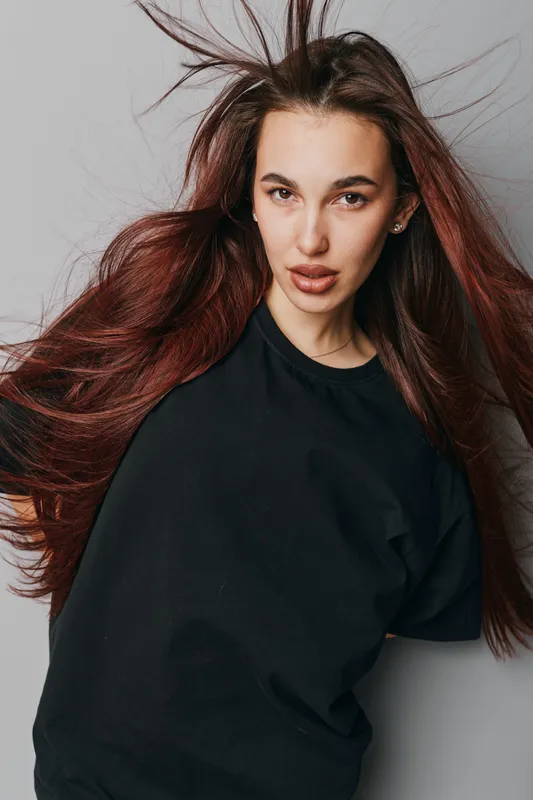 Young woman with long red hair wearing black t shirt posing against gray background, looking at camera with serious expression and natural makeup in studio portrait.