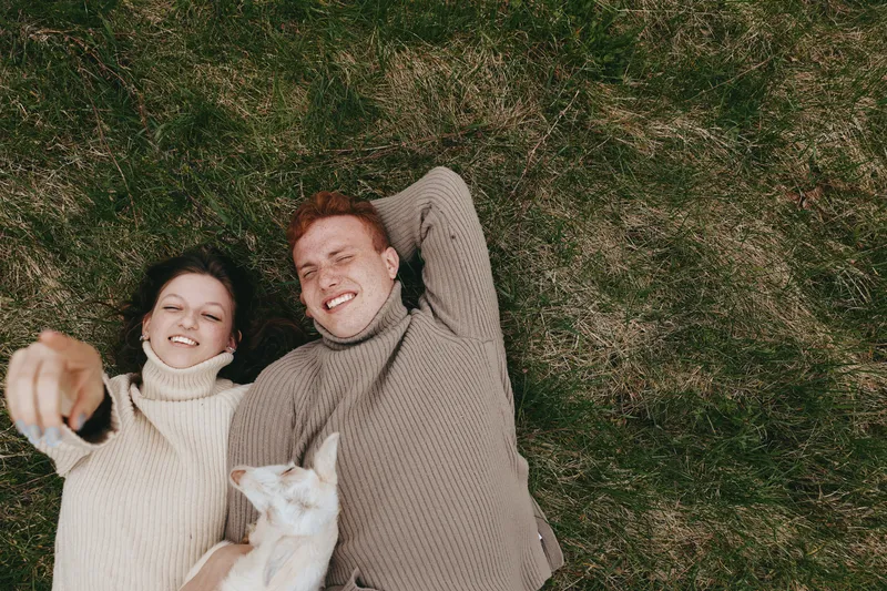 Two smiling people lie on a green lawn in cozy sweaters, a white cat resting on one, capturing a warm lifestyle moment of affection and relaxation outdoors, with shared joy and casual comfort in soft