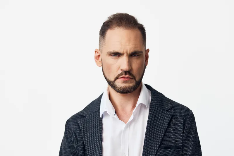 A confident business professional man stands against an isolated white background, exuding focus and determination. He wears a dark textured blazer over a white shirt, ready for a strategic