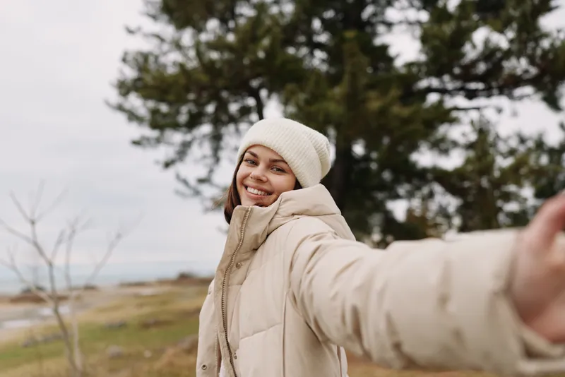 Woman outdoor selfie in a winter coat, smiling at the camera with a landscape of trees and a cloudy sky, portraying a casual outdoor moment