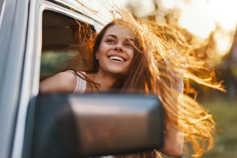 Happy young woman with flowing hair enjoying a summer road trip, embracing freedom and adventure while riding in a car with a picturesque background.