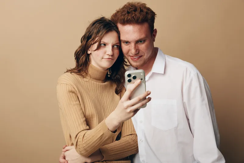 Young couple with red hair and brown hair takes selfie on smartphone. They stand close together against beige background. Concept of modern relationships, technology, and casual lifestyle.