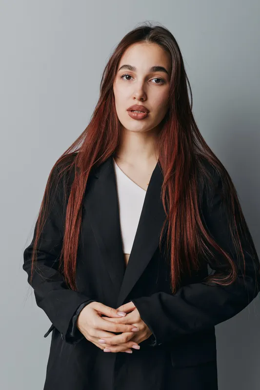 Woman with long hair wearing black blazer and white top standing against plain background looking at camera with serious expression and hands clasped in front of body.