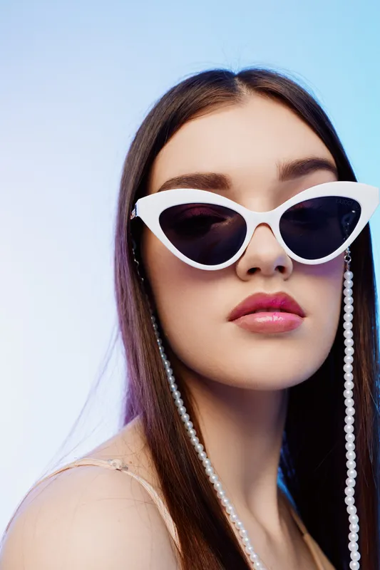 Young woman with long hair wearing white cat eye sunglasses and pearl chain looks confidently at camera. Studio shot on light blue background. Concept of beauty fashion style.