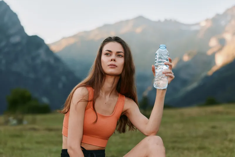 A young woman sits outdoors in a mountain meadow, wearing an orange top and holding a water bottle. She looks toward the horizon, presenting wellness, fitness, and nature in a peaceful scene.