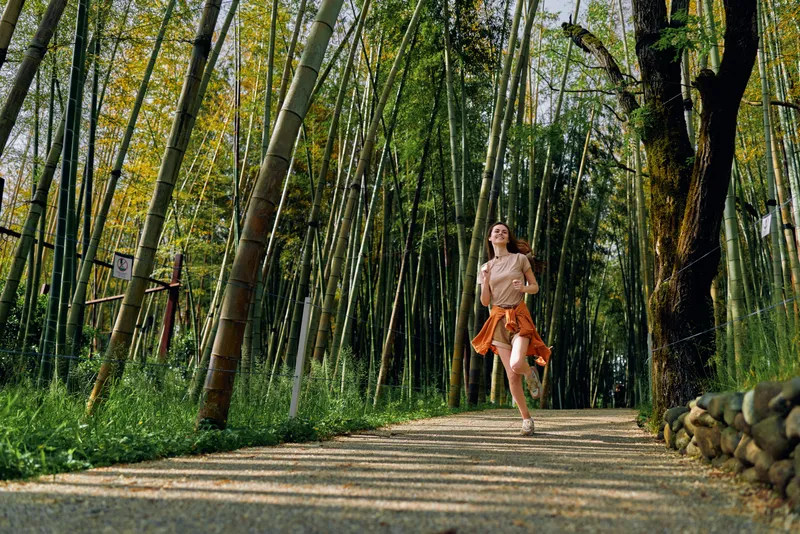 Woman running along a sunlit bamboo forest path, casual summer outfit with tied jacket, enjoying outdoor fitness and jogging on a gravel trail among tall green stalks and dappled light.