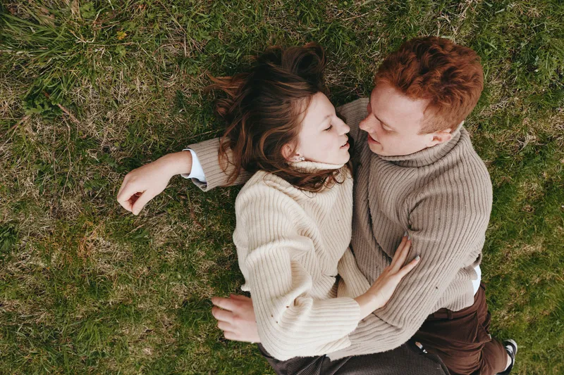 A couple lies on grass outdoors, embracing in a cozy scene. Soft sweaters and relaxed mood capture a tangible lifestyle moment of affection, intimacy, and natural connection.