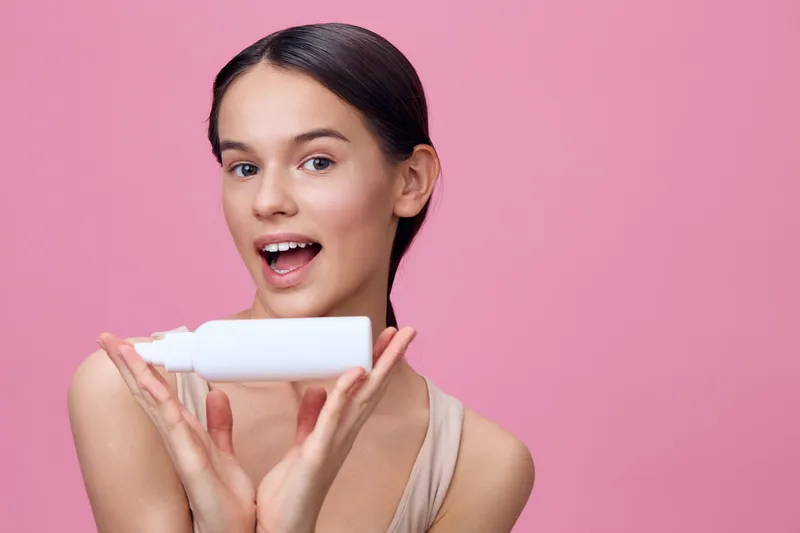 Young woman with a joyful expression holding a white skincare product against a pink background, showcasing beauty and self care concepts in a vibrant and trendy setting