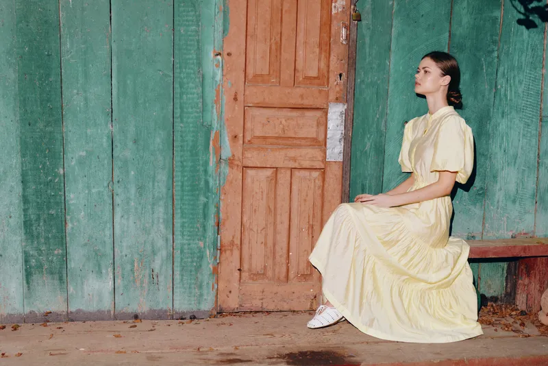 Portrait of a woman sitting on a bench beside a door and green wall, wearing a yellow dress, exuding calm charm and rustic outdoor atmosphere.