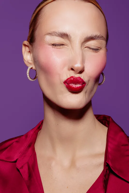 Close-up of a woman with red lipstick and hoop earrings making a kiss face. She wears a red shirt and poses against a purple background. Concept of beauty, makeup, and expression.