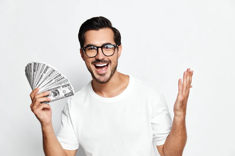 Excited cheerful young man in white t-shirt holding fan of dollar bills, smiling happily at the camera against plain solid white background. Lifestyle concept