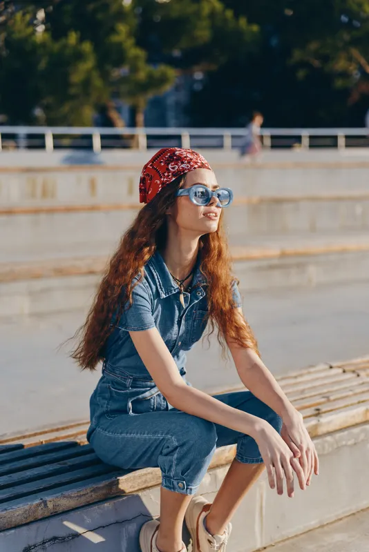 young woman sitting outdoors wearing sunglasses and a red bandana, dressed in blue denim under warm sunlight on a casual summer day