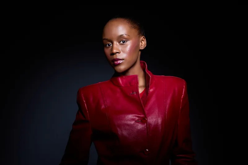 Confident African woman with dark skin wearing a red leather jacket poses against dark background. Studio portrait with dramatic lighting highlights elegance and fashion style.