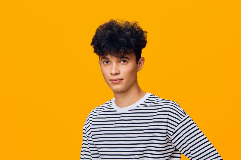 young man with curly hair wearing white and black striped shirt posing against bright orange background with confident and thoughtful expression