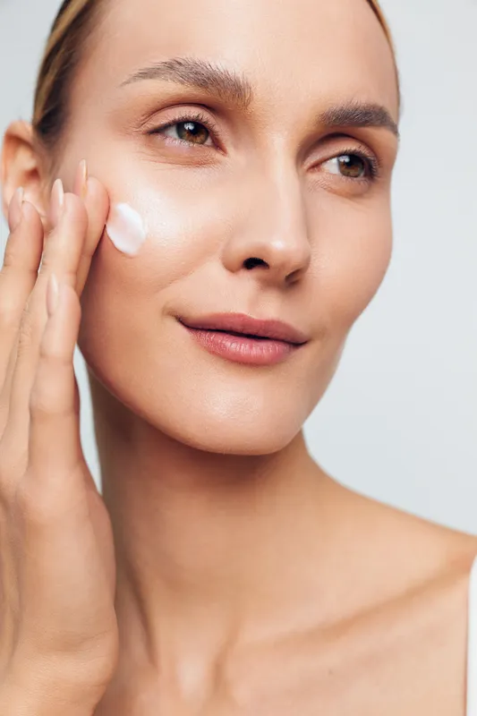 Young woman applies moisturizing cream on her cheek with fingers closeup natural skin care routine isolated on light background beauty concept for healthy face and fresh complexion.