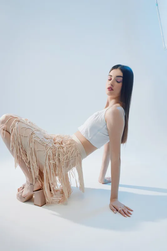 Young woman in fashionable beige fringe skirt and white top poses with closed eyes in bright studio. Concept of beauty, fashion, and modern style.