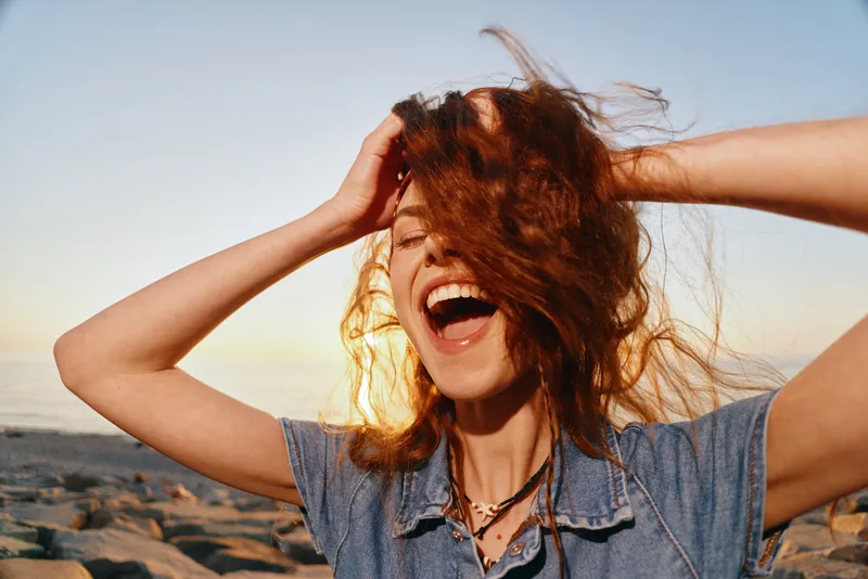 Lifestyle woman with modern boho-western denim street style laughing joyfully outdoors at sunset with natural red hair and carefree energy, film color effect enhancing warmth.