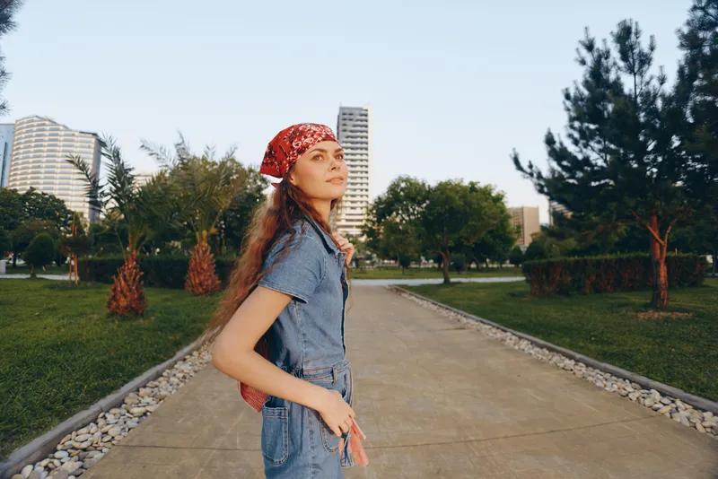 Lifestyle woman wearing modern boho-western denim street style with a red bandana, standing confidently outdoors in urban park under natural sunlight with film color effect.