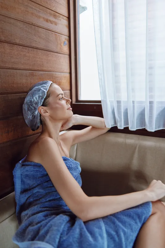 Relaxed woman wrapped in blue towel wearing a shower cap, sitting indoors by the window with soft natural light and wooden wall background, enjoying peaceful moment.