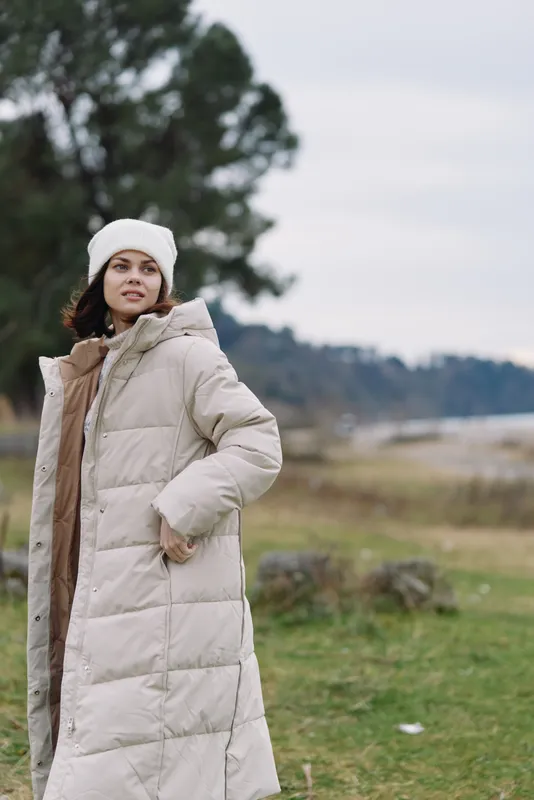 Winter fashion scene featuring a woman in a long beige puffer coat and knit hat, standing in an open field with soft trees in the distance and a calm, cloudy sky