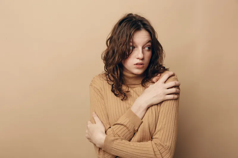 A worried young woman with curly hair wearing a brown sweater looks to the side with crossed arms. The neutral beige background emphasizes her anxious and thoughtful mood.