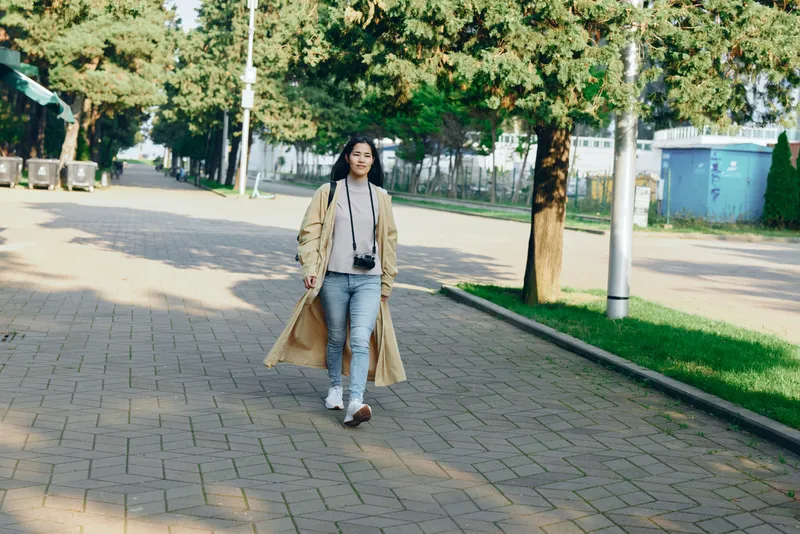 A stylish woman walks along a park path wearing a long coat and carrying a camera, exploring outdoors with casual fashion under daylight and urban mood