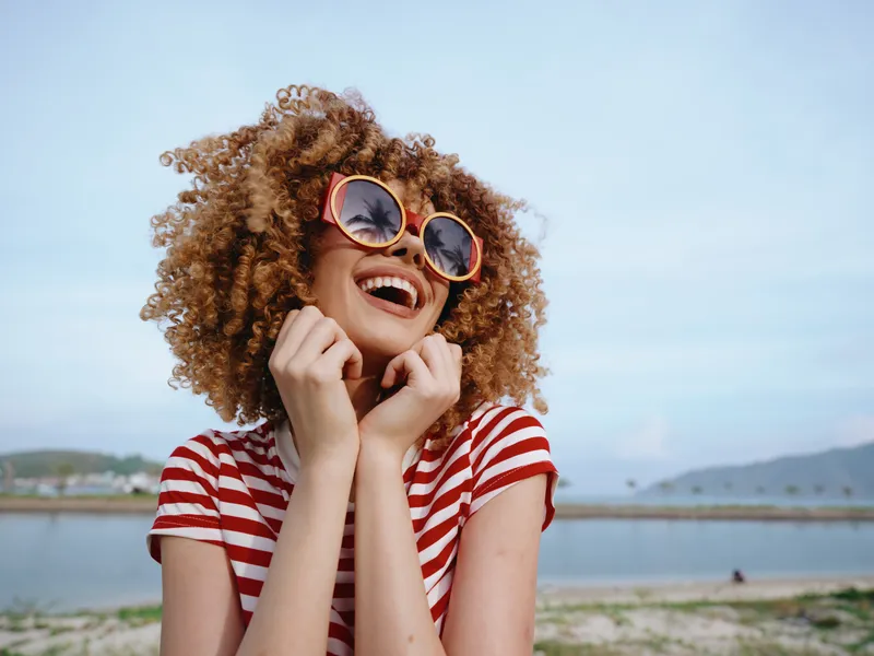 Joyful young woman with curly hair wearing striped shirt and round sunglasses smiles brightly at the beach, capturing a carefree summer moment under clear blue skies.