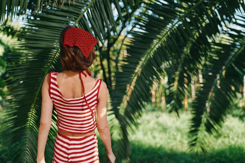 A young woman stands with her back to the camera in a sunny tropical garden. She wears a red and white striped top and a matching red hat, enjoying a bright outdoor moment.