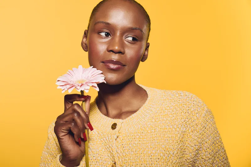 Portrait of a dark skinned African woman holding a pink flower near her face against yellow background. Concept of natural beauty, skincare, and self-care with warm lighting and cozy clothes.