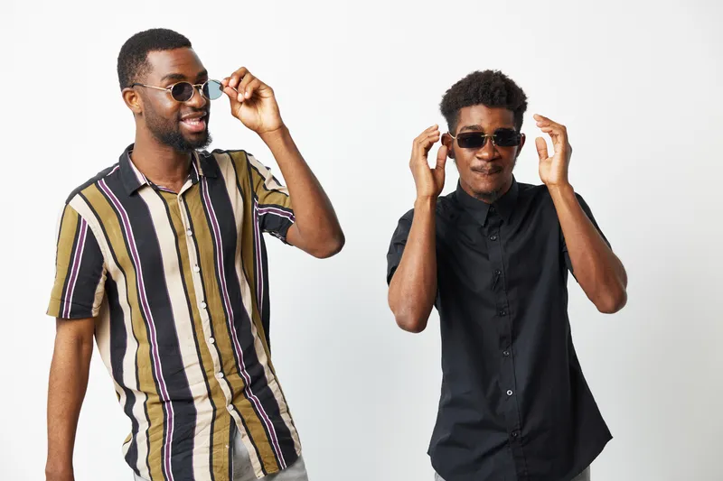 Two stylish African American men wearing sunglasses showcase their cool vibes, embodying confidence and fashion in a modern, upbeat atmosphere.