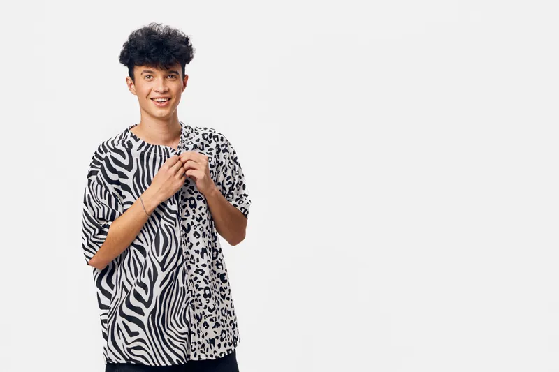 young man in patterned casual t-shirt stands smiling against white background, expressing joy and style with mixed animal print and curly hair