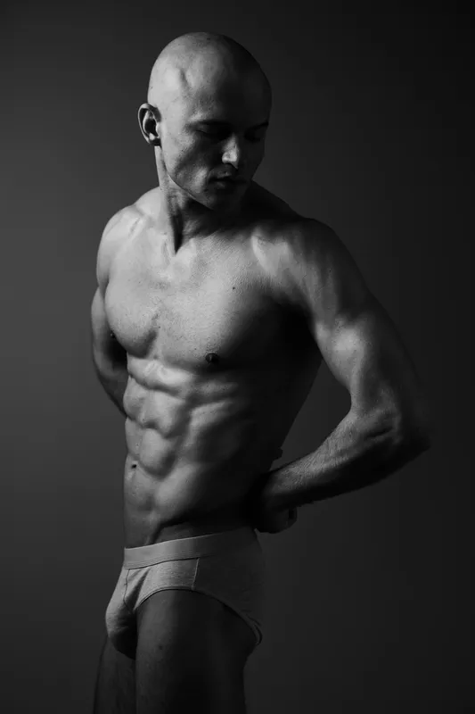 Black and white image of a muscular man showcasing his physique with a confident pose, emphasizing strength and determination in a minimalist setting.