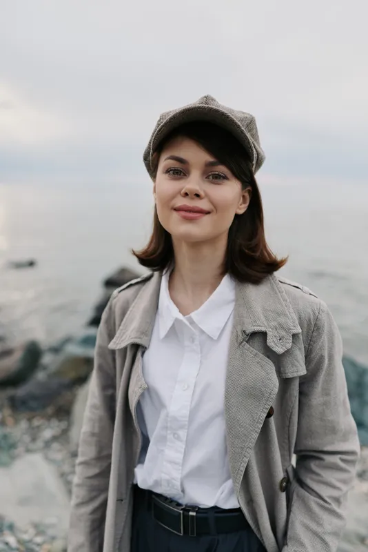 Young woman with short dark hair wearing a grey coat and hat stands outdoors near rocky shore. She smiles gently with calm ocean and cloudy sky in background. Casual autumn fashion.