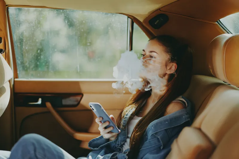 Young woman relaxing in car, exhaling vapor while using smartphone, showcasing modern lifestyle and casual fashion in soft, natural light.