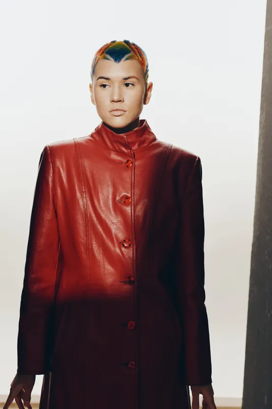 woman with short colorful hair wearing a red leather coat standing against a bright background with soft shadows, modern fashion style.
