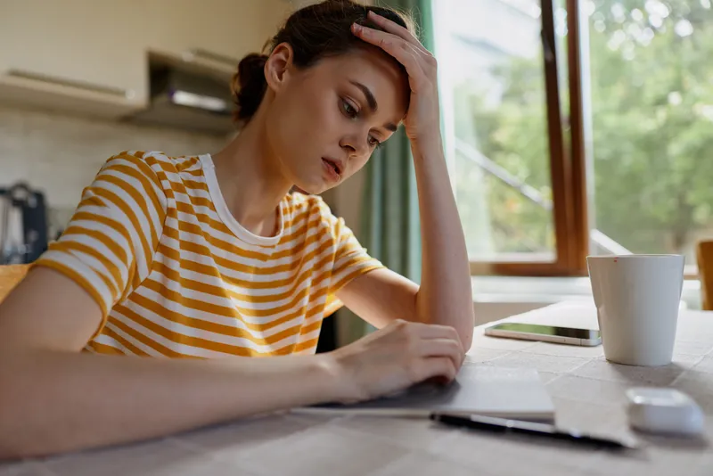 A pensive adult woman sits at a home desk with papers scattered around, resting her forehead in frustration, a mug nearby in a cozy kitchen light.