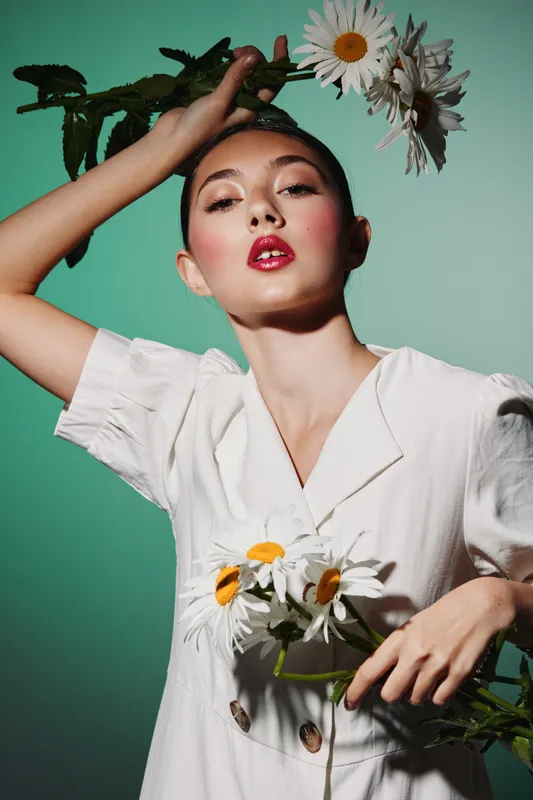 Stylish beauty with glossy red lips holds fresh daisy flowers near face, wearing white outfit against soft green background. Concept of fashion, creativity, summer, and elegance.