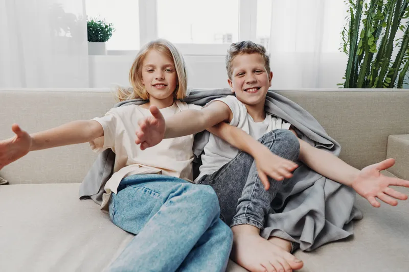 Two happy children sitting on a couch with open arms and smiling faces, enjoying cozy time at home in a bright living room setting.