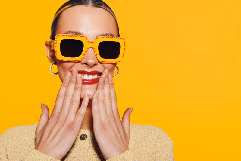 Smiling woman with orange sunglasses and red lipstick covers mouth with hands on bright yellow background. Concept of happiness, fashion, and summer style with vibrant colors and accessories.