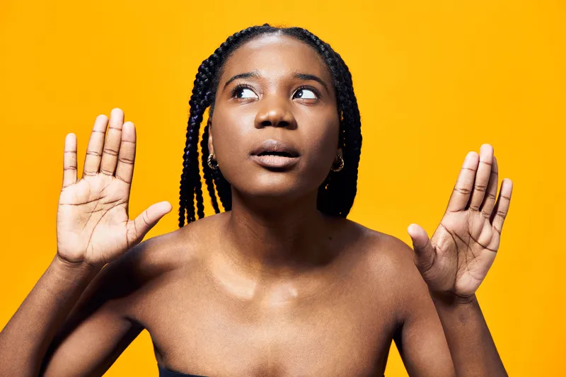 Expressive Black woman with braided hair against a vibrant yellow background, conveying a sense of surprise and curiosity while wearing a strapless top
