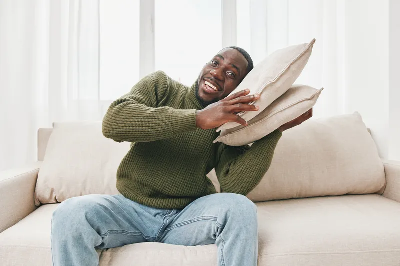 Happy young African American man enjoying his cozy living room with pillows, wearing green sweater and denim jeans, exuding warmth and comfort
