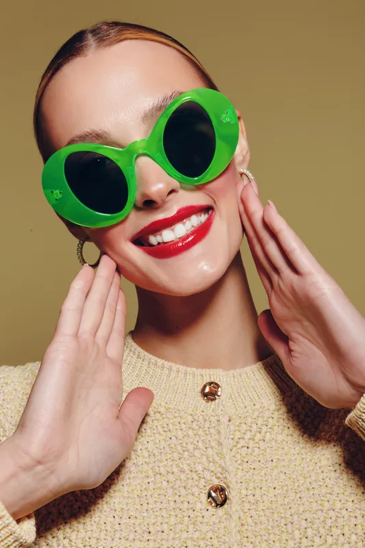 Smiling woman with bright red lipstick wears large green sunglasses and beige sweater, posing with hands near face on neutral background. Concept of fashion, style, and beauty.