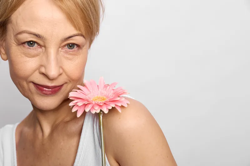 Mature woman closeup portrait with pink gerbera daisy flowers, expression and soft beauty lighting on white background, concept of skincare, wellness, aging.
