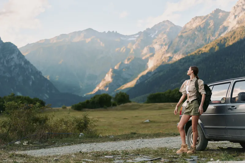 Smiling woman in hiking attire stands beside a car in a mountainous valley, ready for a trek. Outdoor lifestyle scene with rugged terrain and clear sky.