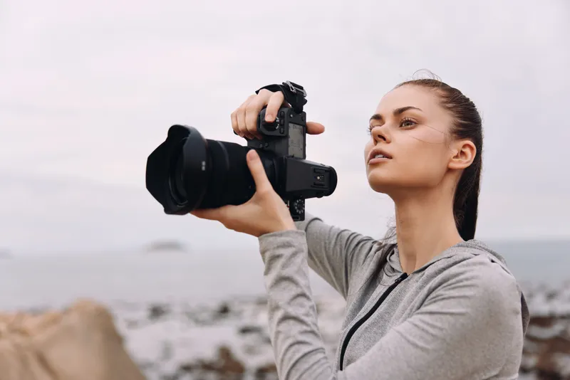 Young woman capturing nature with a camera at the beach, showcasing passion for photography, stylish in a grey hoodie, with a serene sea backdrop