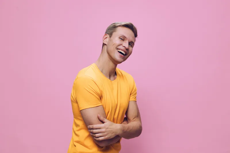 Happy smiling man wearing yellow t-shirt standing crossed arms against pastel pink background joyful positive emotion concept