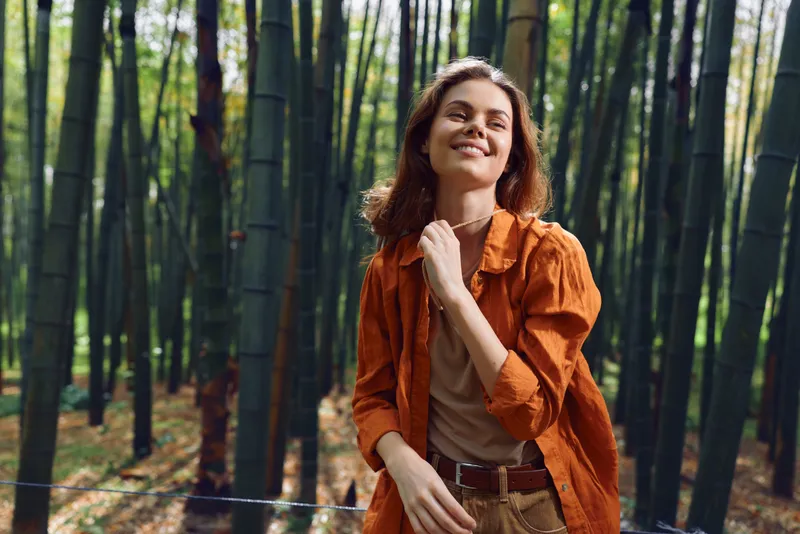 Woman portrait smiling in bamboo forest, nature outdoors and casual fashion. Happy relaxed young adult enjoying sunlight among tall bamboo stalks for eco travel and leisure.