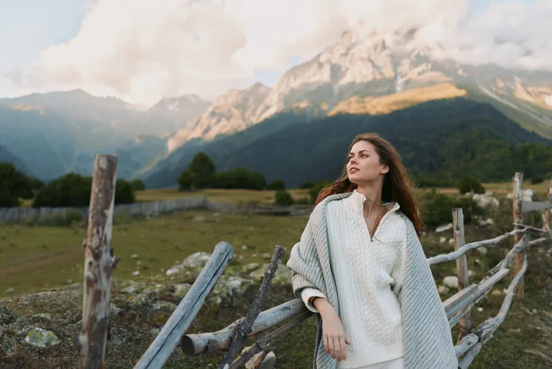 Woman in a white coat stands near a wooden fence in a mountain meadow, enjoying a calm outdoor lifestyle amid expansive nature and rugged scenery.