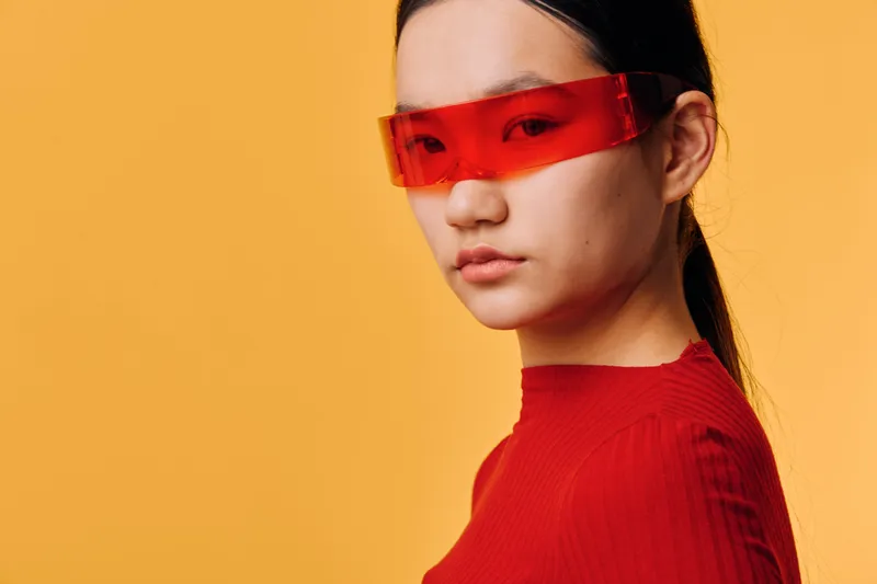 Woman with red futuristic glasses and red sweater posing against orange background. Modern fashion, portrait with serious expression and sleek hairstyle in studio lighting.